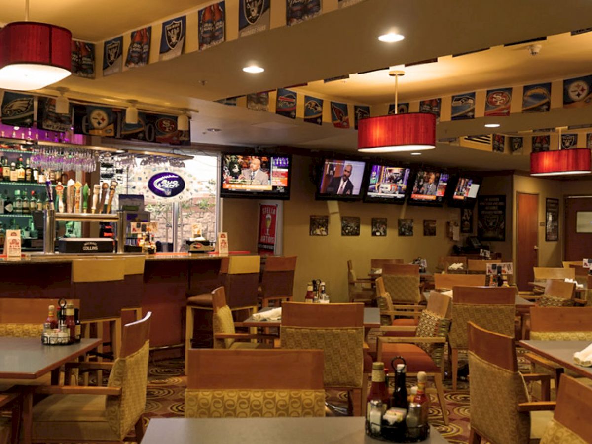 The image shows a cozy restaurant bar with tables and chairs, a well-stocked bar, multiple TVs, and pendant lights, creating a welcoming atmosphere.