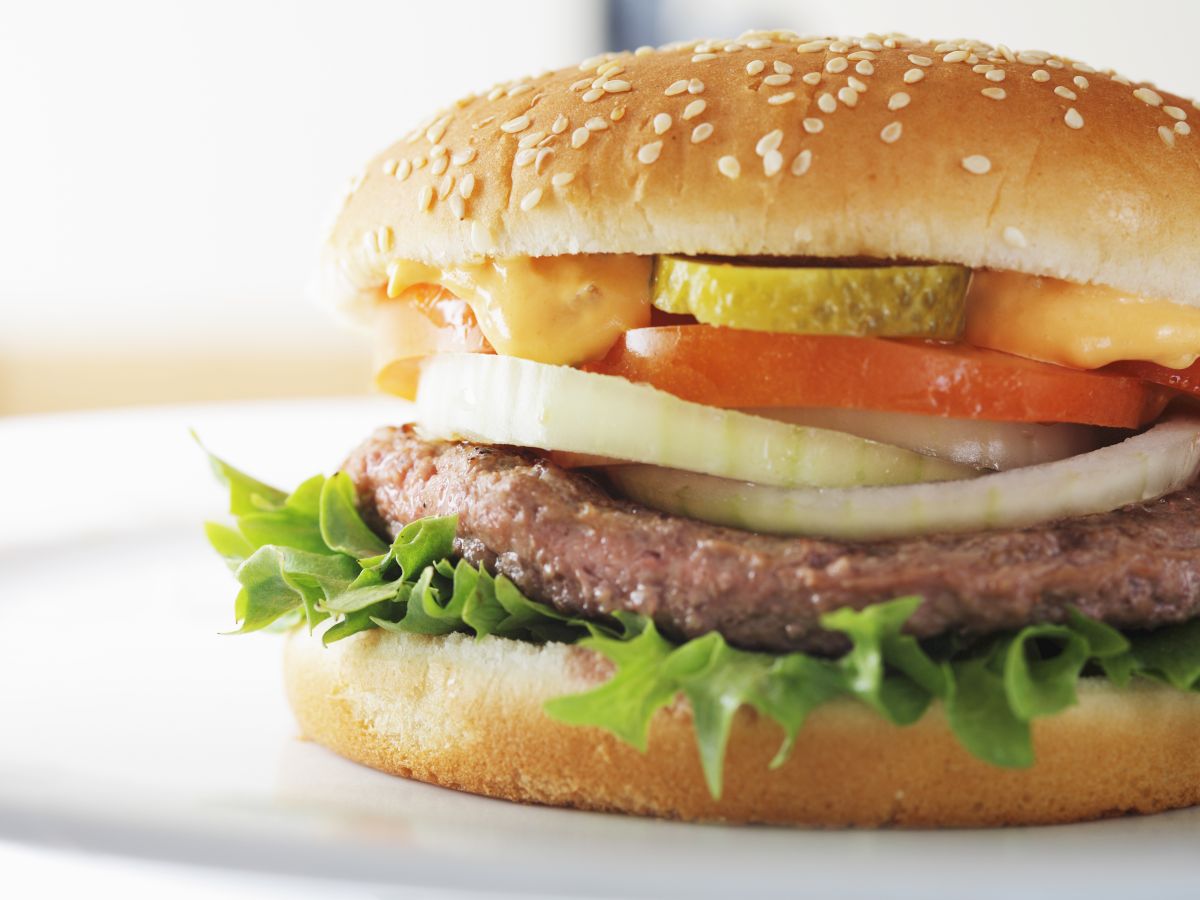 The image shows a close-up of a hamburger with lettuce, a beef patty, onions, tomato, pickles, and sauce inside a sesame seed bun.