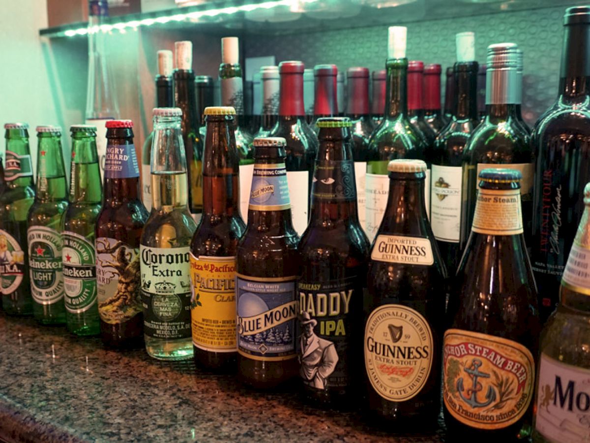 The image shows a variety of beer bottles and wine bottles lined up on a countertop with some recognizable brands visible.