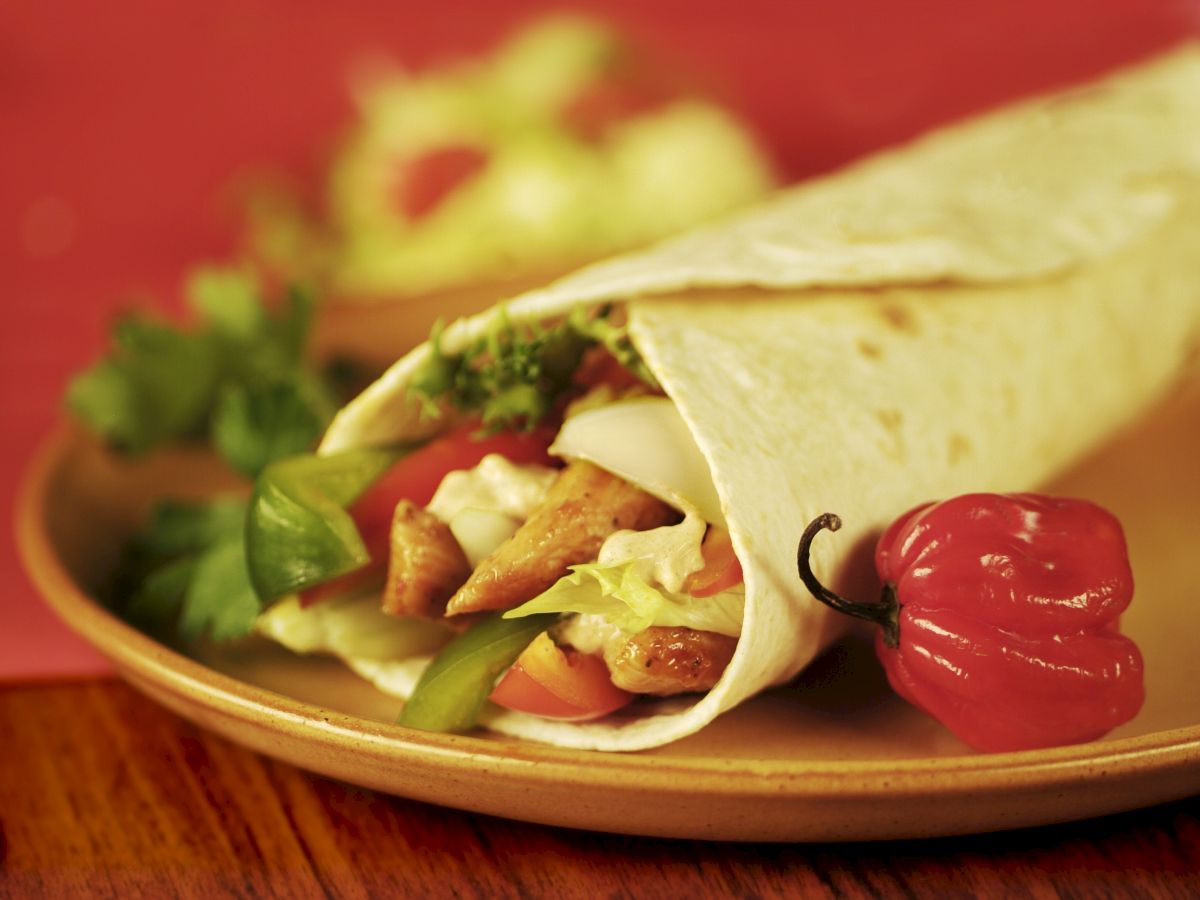 A close-up of a wrapped tortilla with fillings like chicken, lettuce, tomatoes, and bell peppers is served on a plate with a red chili pepper.