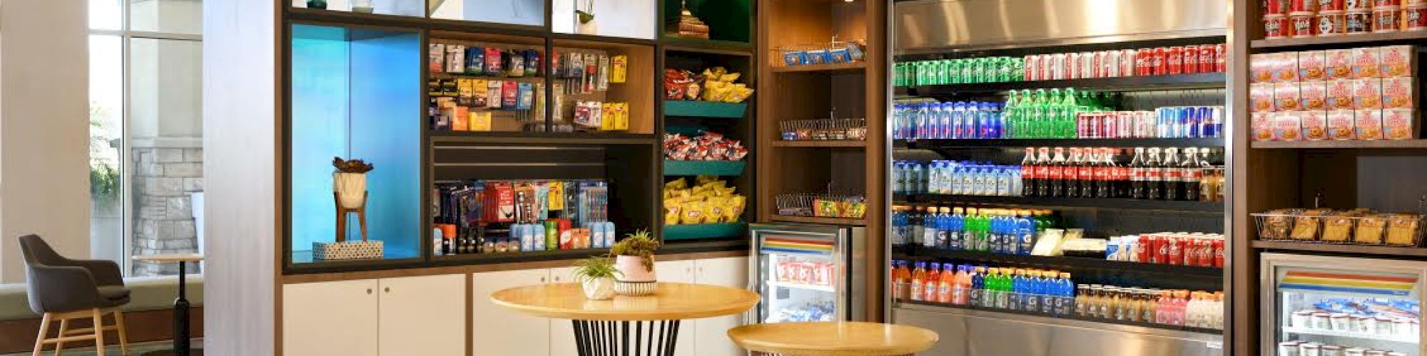 A modern convenience store with shelves of snacks and beverages, a refrigerated section, and three round tables with seats in the foreground.