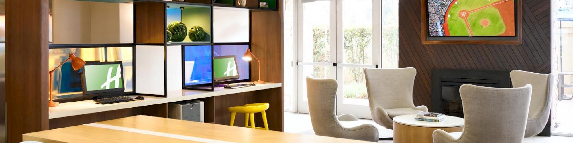 A modern communal workspace with a long table, blue chairs, and a shelving unit, adjacent to a lounge area with armchairs and a TV.