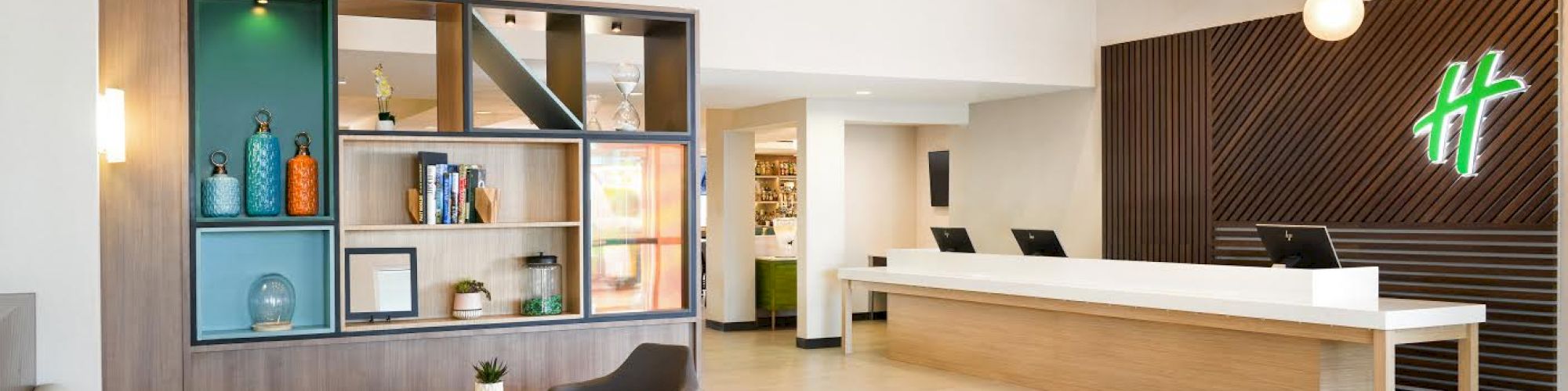 The image shows a modern hotel lobby with a reception desk, seating area, shelves with decor, and hanging lights. The decor is minimalist and inviting.