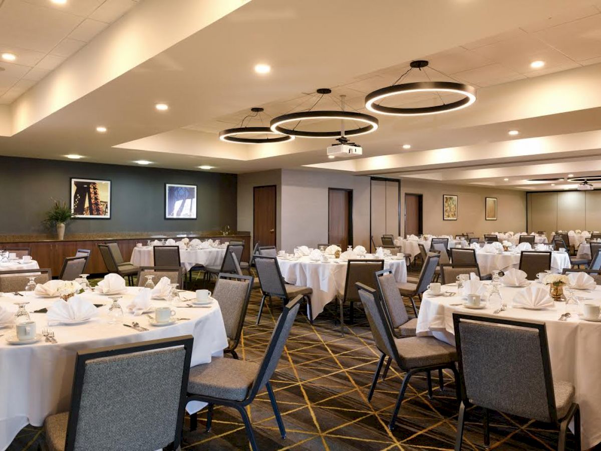 The image shows a banquet hall set for an event, with round tables, chairs, white tablecloths, and modern lighting fixtures in a spacious room.