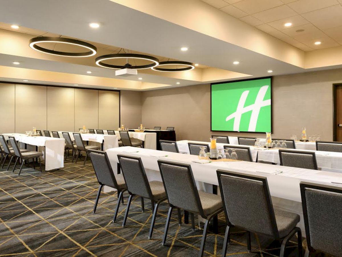 This image shows a conference room set up with tables and chairs, featuring a large screen displaying a green "H" logo.