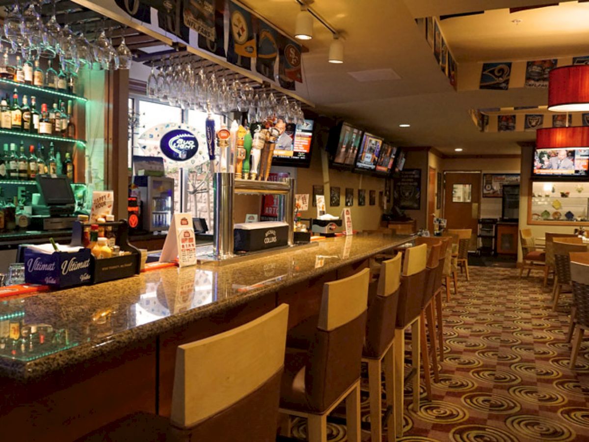 The image shows an empty bar with a long counter, bar stools, various alcoholic beverages on shelves, and multiple TV screens.