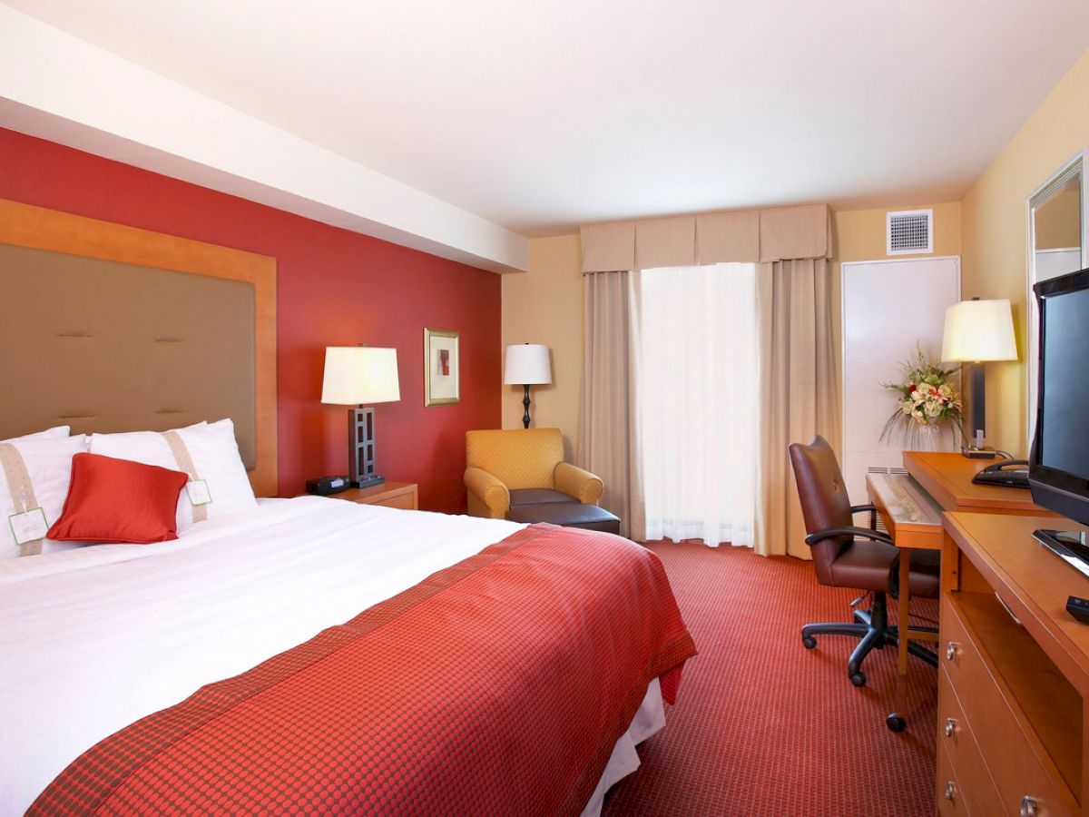 A hotel room with a large bed, red accents, two lamps, an armchair, a desk with a chair, a TV, and a window with curtains.