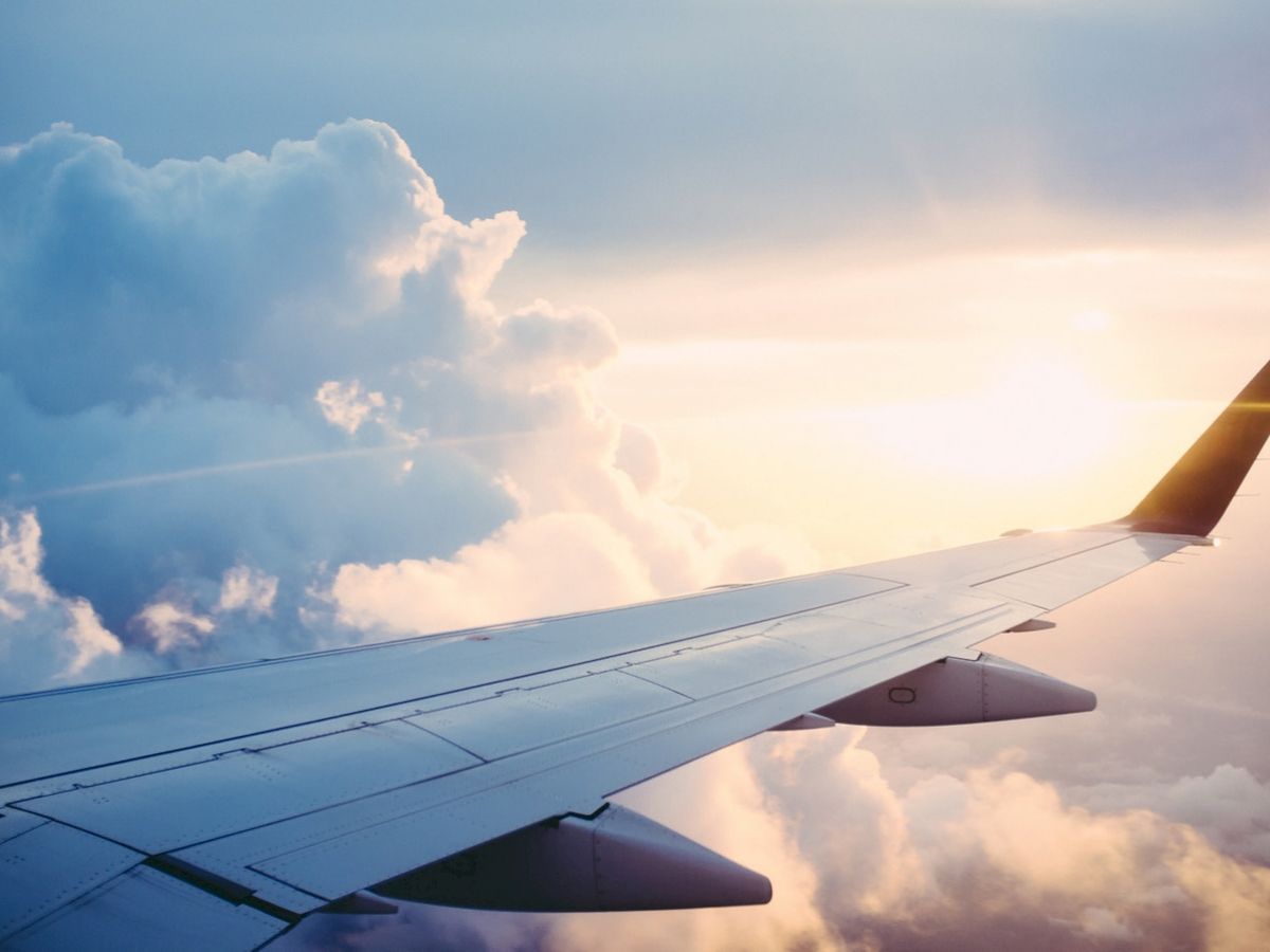 The image shows an airplane wing against a backdrop of a blue sky with clouds and a setting or rising sun in the background.