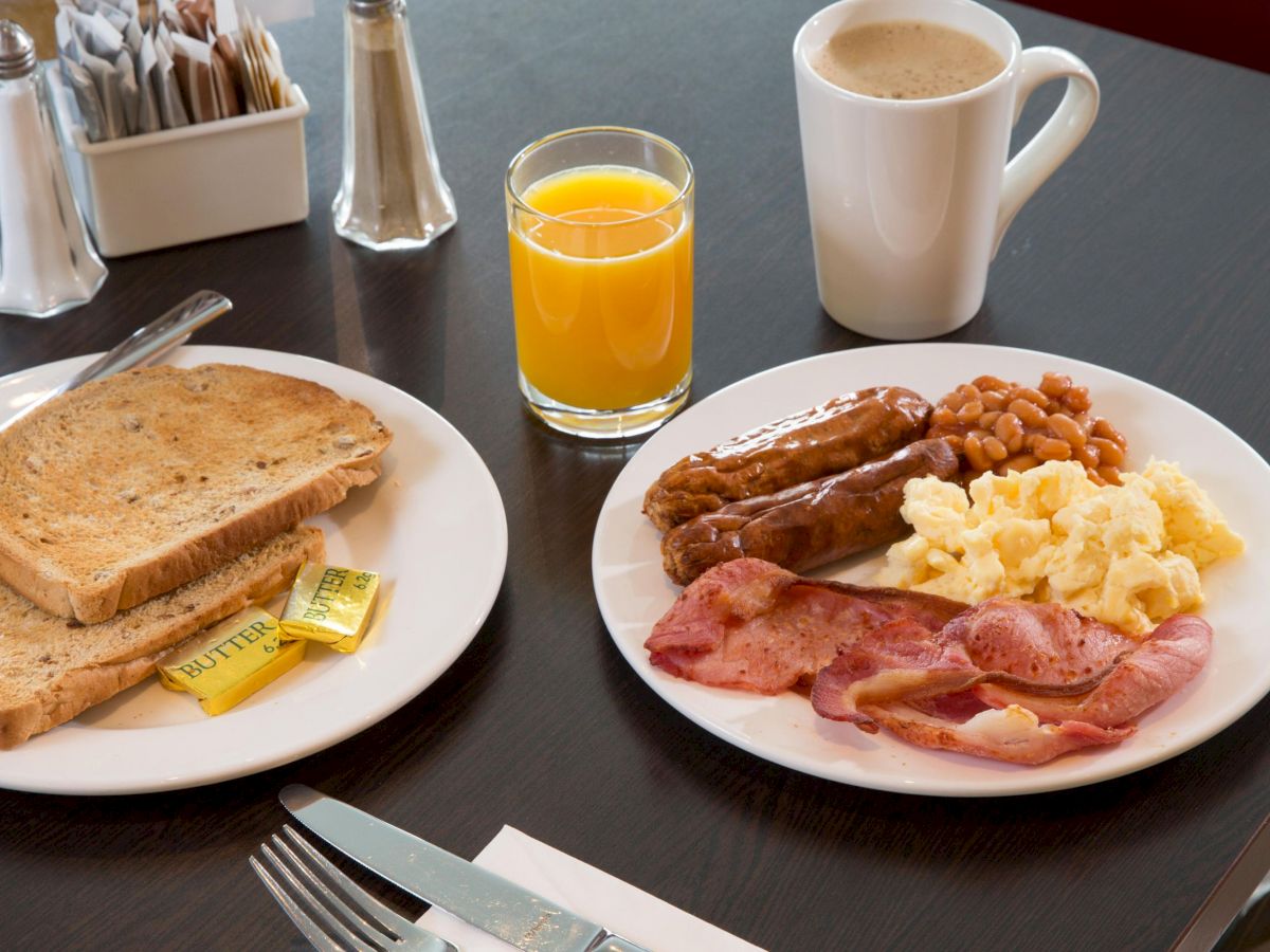 A breakfast setup with toast, butter, bacon, sausages, scrambled eggs, baked beans, orange juice, and coffee on a table.