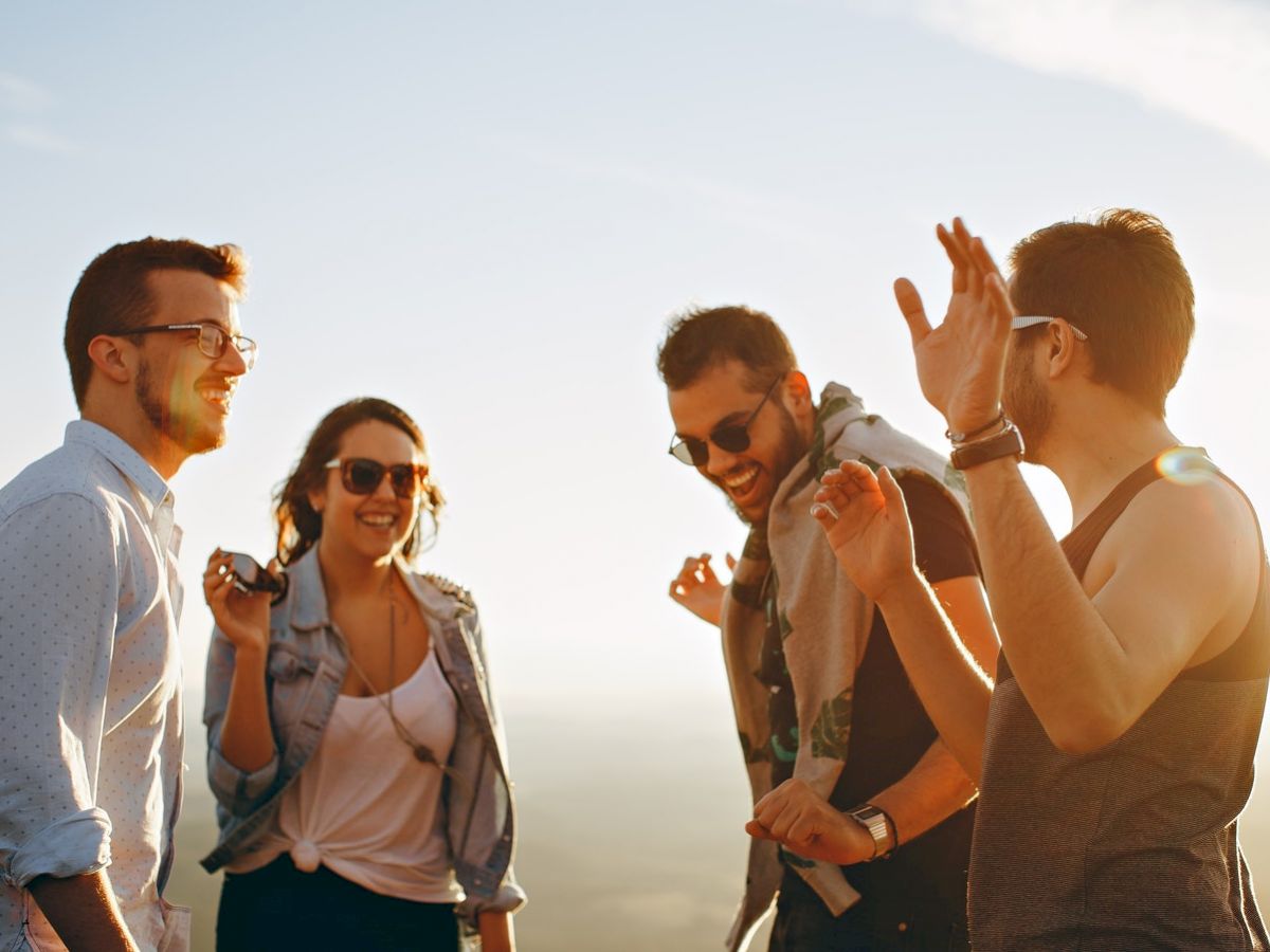 Four people are outdoors, laughing and enjoying each other's company under a bright sky, suggesting a casual and joyful moment.