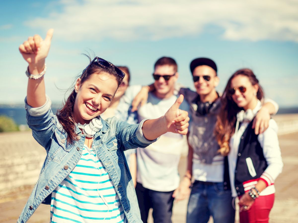 A group of friends enjoying a sunny day outdoors, with one girl in the front giving a thumbs-up and smiling, while the others pose in the background.