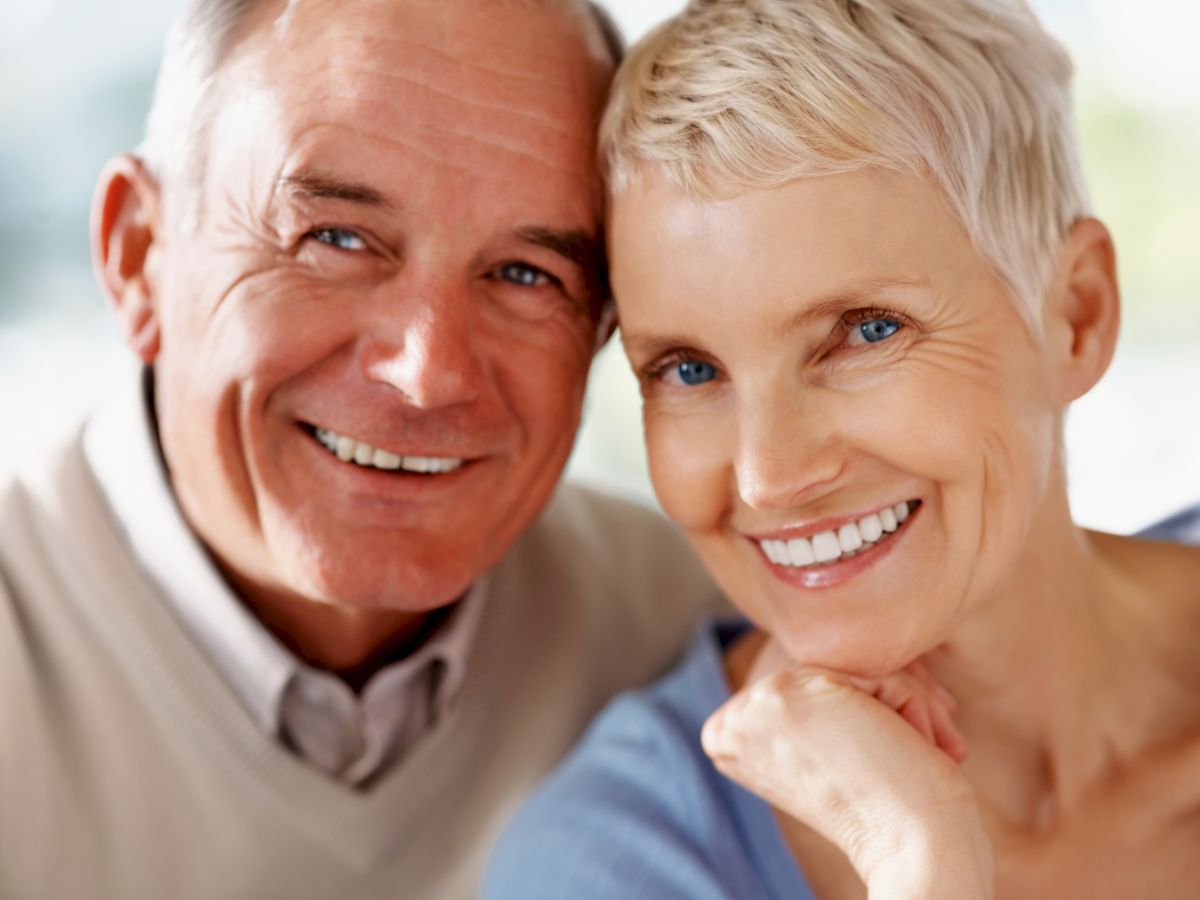 An elderly couple is smiling at the camera, appearing happy and content. They are dressed casually and seated closely together indoors.