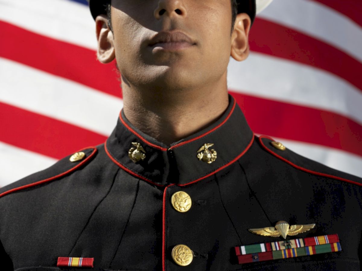 A person in military uniform stands in front of an American flag. The uniform features medals and insignia, with a white cap indicating possible naval service.