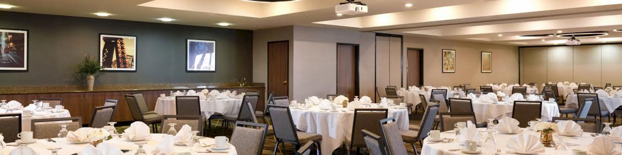 A banquet hall with round tables set with white tablecloths, napkins, and chairs, modern lighting fixtures, and framed artwork on the walls.