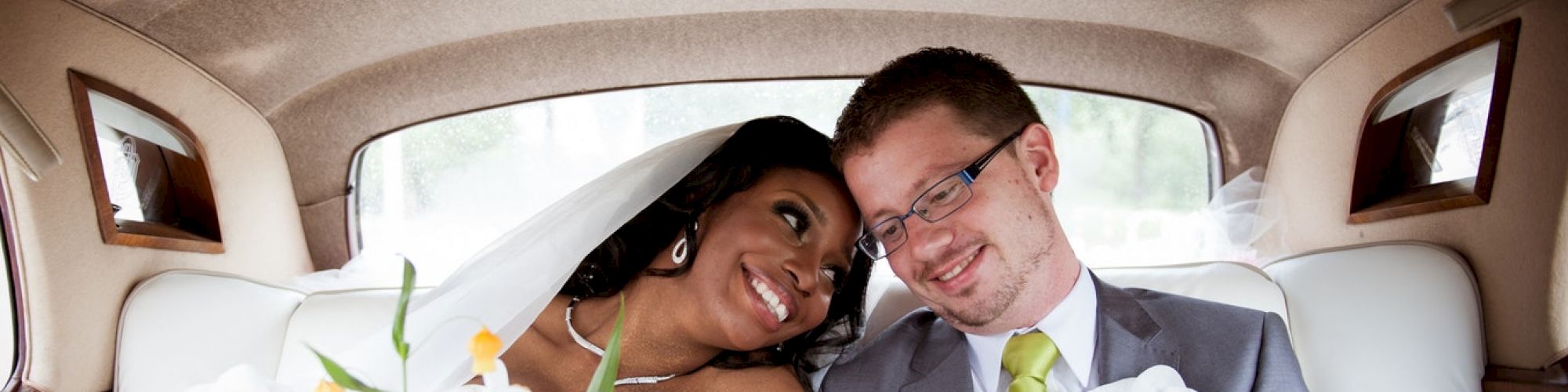 A happy couple, dressed in wedding attire, is sitting closely together in the back of a car, smiling warmly at each other.