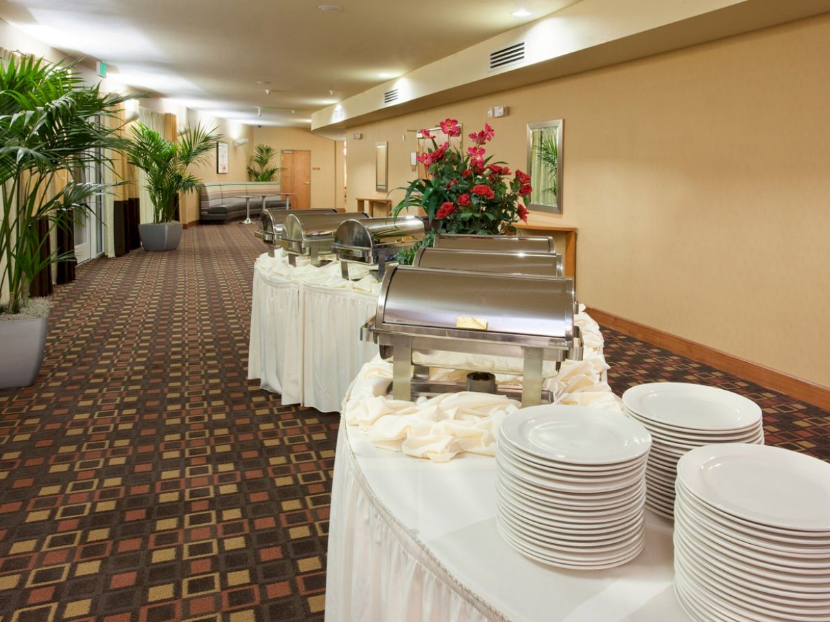 A buffet setup in a hallway with chafing dishes, stacks of plates, napkins, and floral decor, alongside potted plants under a well-lit ceiling.