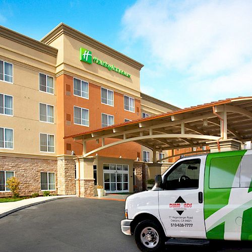 The image shows the exterior of a Holiday Inn hotel with a canopy entrance and a van parked in front, featuring company branding.