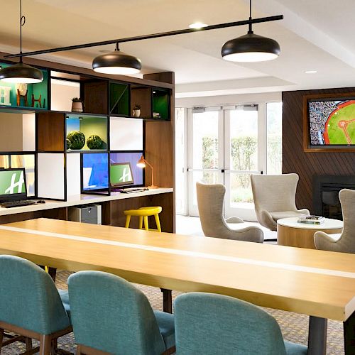 The image shows a modern lounge area with a long wooden table and turquoise chairs, a shelving unit with decor, and a TV displaying a baseball field.