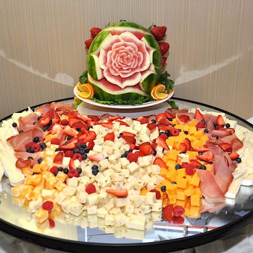 A large platter with various cheeses, deli meats, and fruit, topped with a watermelon carved into a flower, is displayed on a reflective surface.