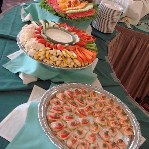 A buffet table with assorted food items, including a vegetable platter with dip, fruit, and appetizers, all elegantly arranged on teal tablecloths.