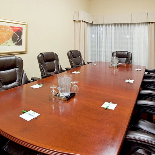 A conference room with a long wooden table, eight leather chairs, water pitchers, glasses, small notepads, pens, and framed art on the wall.