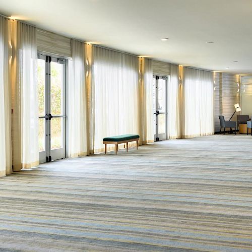 The image shows a spacious lounge area with striped carpeting, large windows with white curtains, a bench, and seating arranged in the background.