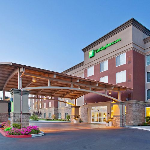 A modern hotel exterior with a covered entrance, marked by landscaped greenery and the hotel's bright signage, is shown under the evening sky.