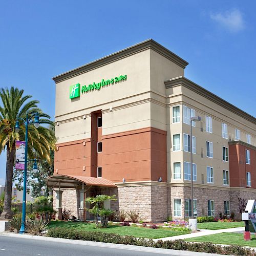 The image shows a multi-story building of a Holiday Inn hotel with a well-maintained exterior and surrounding greenery under a clear blue sky.