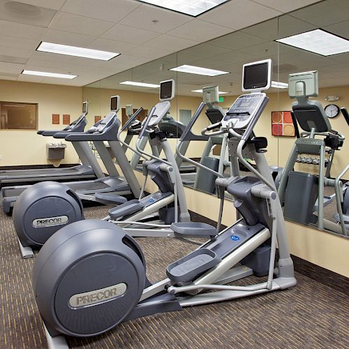 A gym room with various exercise equipment, including treadmills and elliptical machines, with a large mirror on the wall.