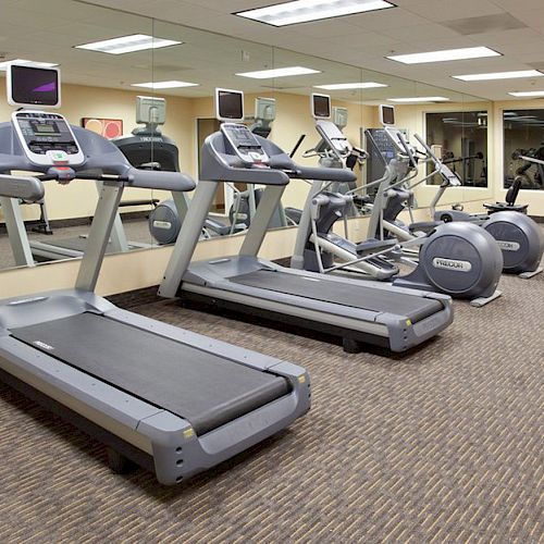 This image shows a gym with treadmills, ellipticals, and exercise bikes lined up in front of mirrors and TVs on the wall.