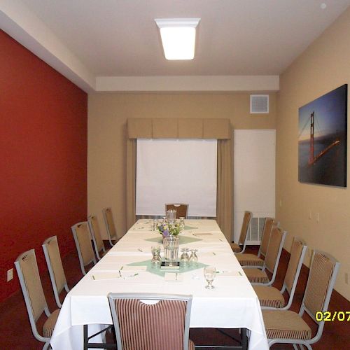 A conference room with a long rectangular table, white tablecloth, surrounded by chairs. Walls have paintings, and the room's lighting is from the ceiling.