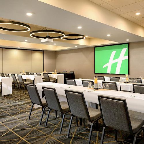 A conference room with long tables, chairs, a projector screen displaying a green logo, and circular light fixtures on the ceiling.