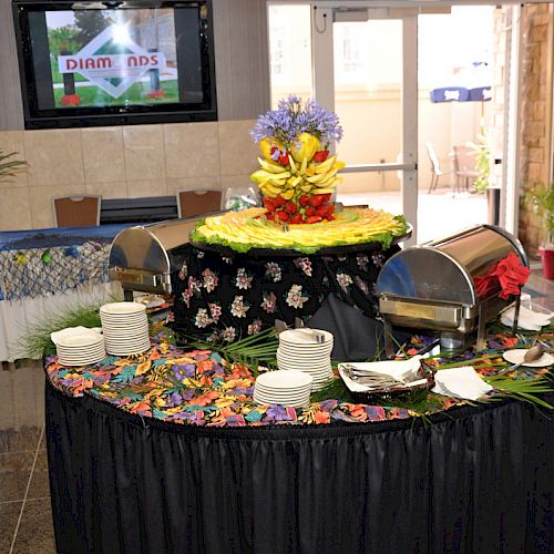 A buffet setup featuring chafing dishes, plates, and colorful decorations, with a flower arrangement and a platter of cut fruit at the center.