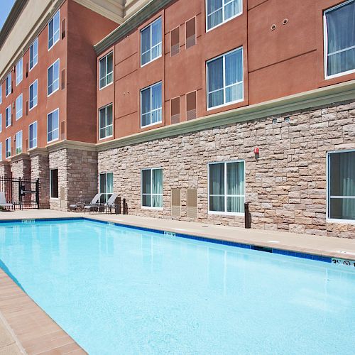 This image shows an outdoor swimming pool adjacent to a multi-story building with stone lower walls and numerous windows, likely part of a hotel.