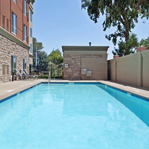 An outdoor swimming pool with clear water next to a building, surrounded by a wall, with trees in the background and safety signage visible.