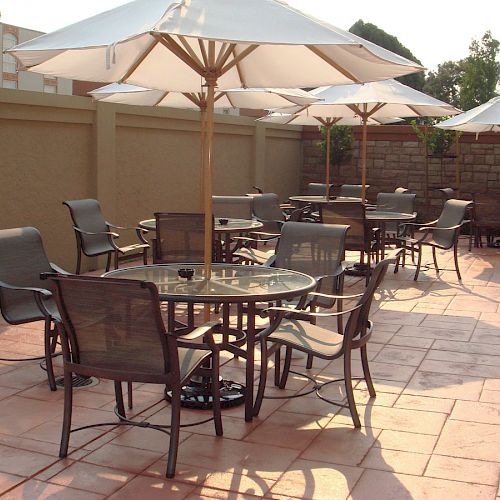 Several outdoor tables with umbrellas and chairs on a tiled patio area are shown, with sunlight casting shadows.