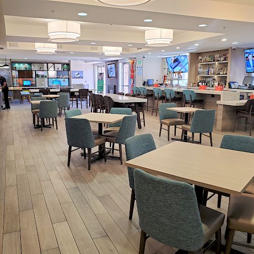 The image shows an empty, modern dining area with tables and chairs, a bar on the right, and TVs on the walls. A few people are visible in the background.