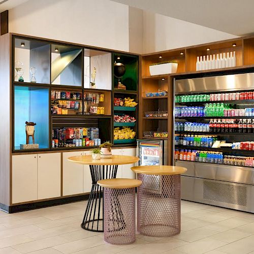 The image shows a small convenience store with shelves of snacks, refrigerators with beverages, and two tables with stools in a well-lit area.