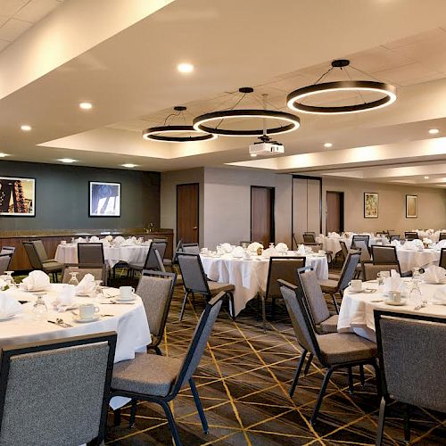 A banquet hall with round tables set for dining, featuring white tablecloths, napkins, and various place settings, under modern circular light fixtures.