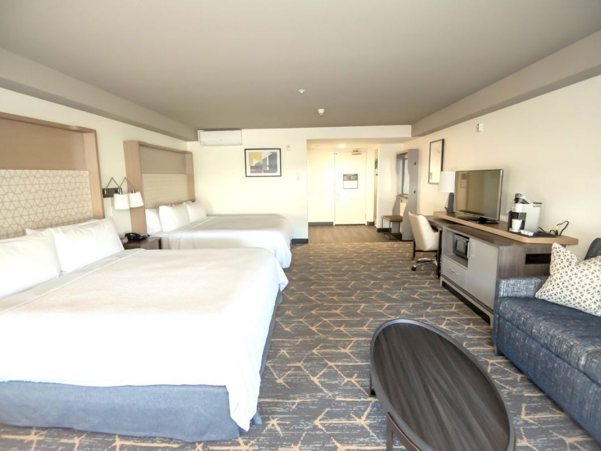 A spacious hotel room with two large beds, a sofa, TV, desk, microwave, mini-fridge, and coffee maker. The room features modern decor and ample lighting.