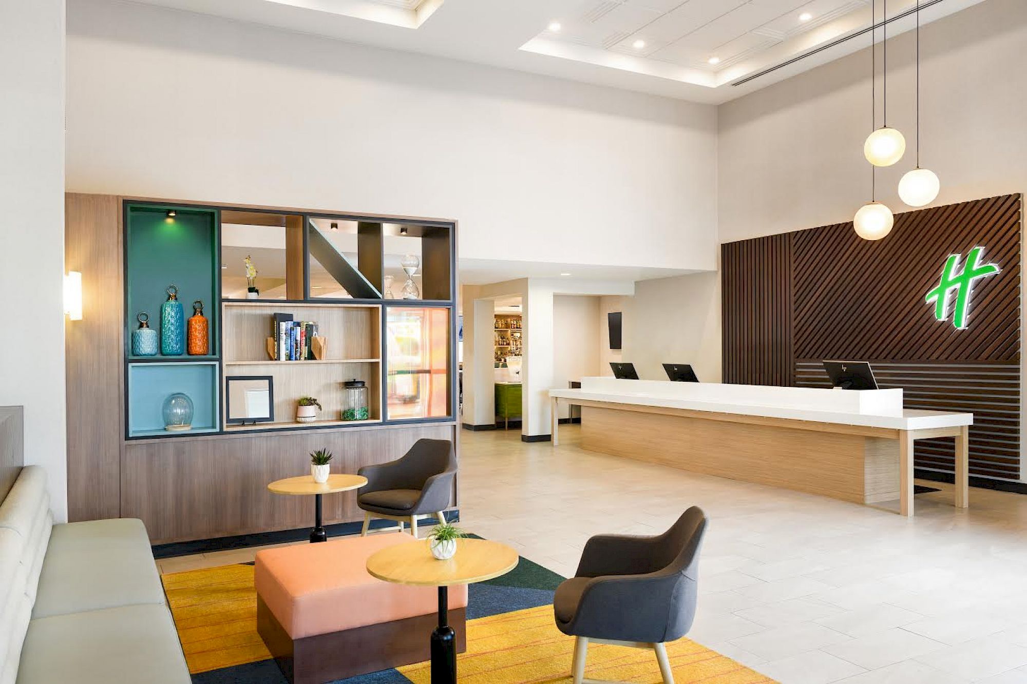 A modern hotel lobby with a reception desk, seating area with chairs and tables, decorative shelving, and a wall-mounted logo.