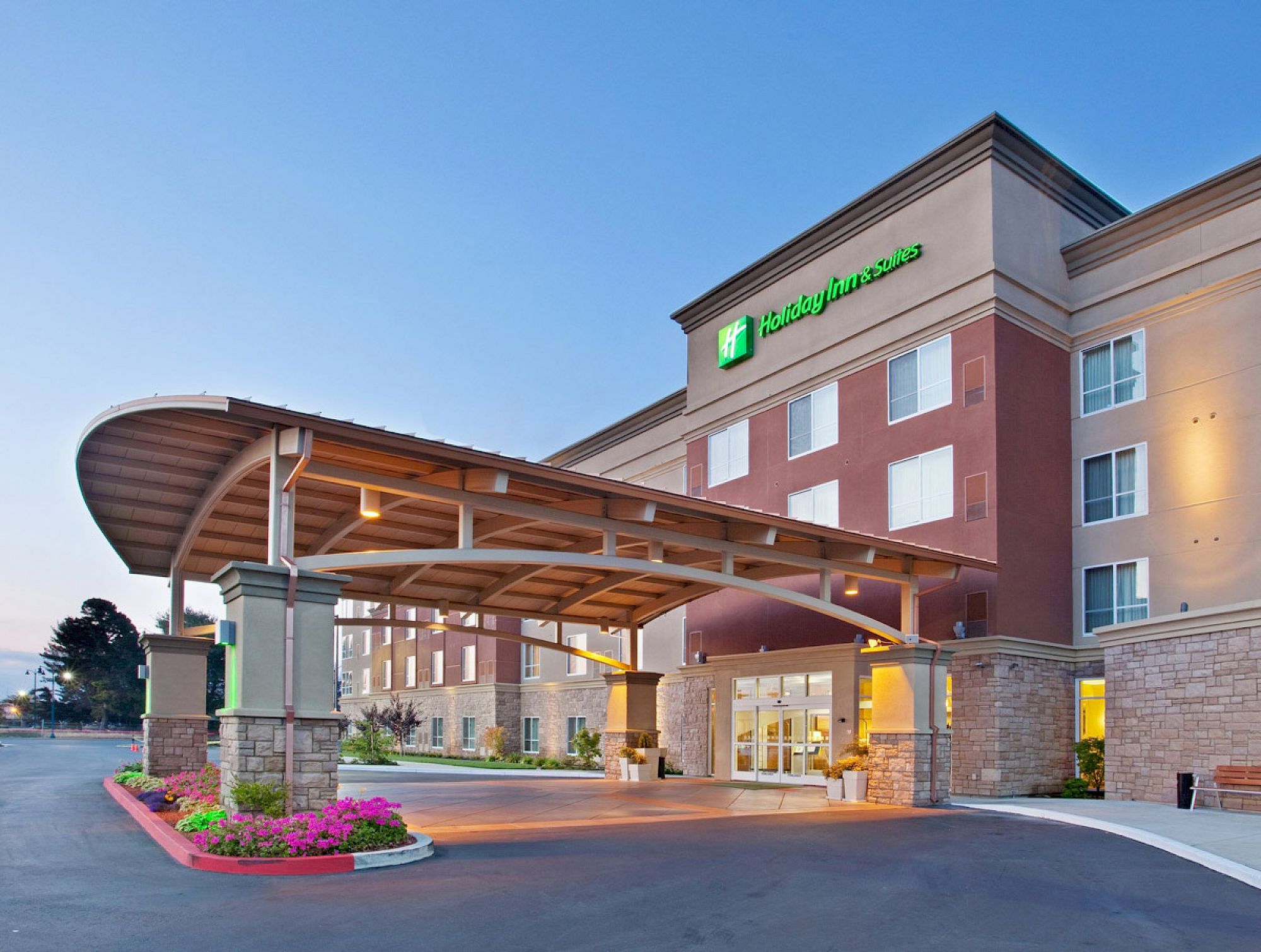 The image shows the exterior of a Holiday Inn hotel, featuring a covered driveway, landscaped entrance, and the brand's logo prominently displayed.