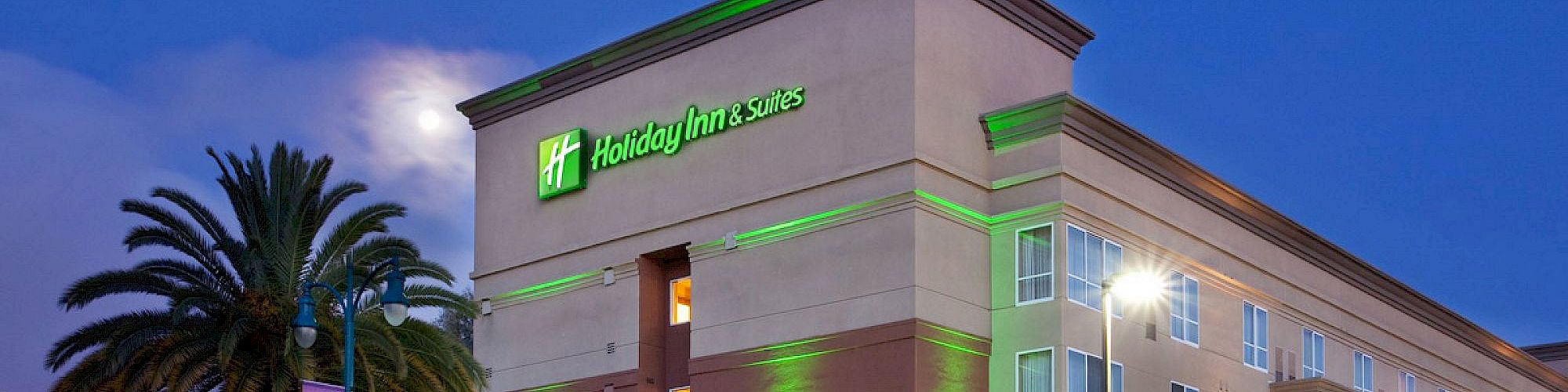A hotel building with green and beige exterior lighting, and palm trees nearby, viewed during twilight.