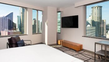 A modern hotel room with large windows showcasing a cityscape, a wall-mounted TV, a chair, and a wooden console table under the TV.