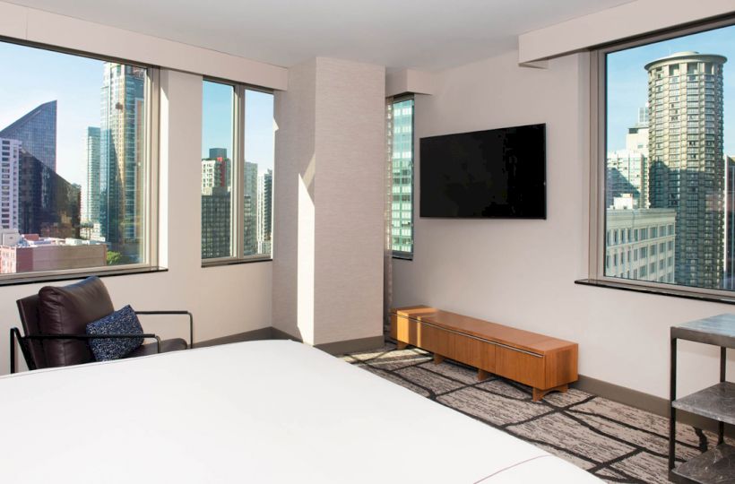 A modern hotel room with large windows showcasing a cityscape, a wall-mounted TV, a chair, and a wooden console table under the TV.