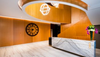 A modern, elegant lobby with a marble reception desk, a large wall clock, wood paneling, spiral staircase, and a floral arrangement.