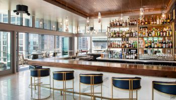 A modern bar features a sleek counter with five stools, shelves stocked with various alcohols, and large windows offering a cityscape view.