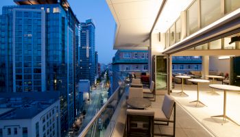 A modern urban balcony at dusk showing city buildings, glass railings, outdoor seating, tables, and large windows under a lit overhang.