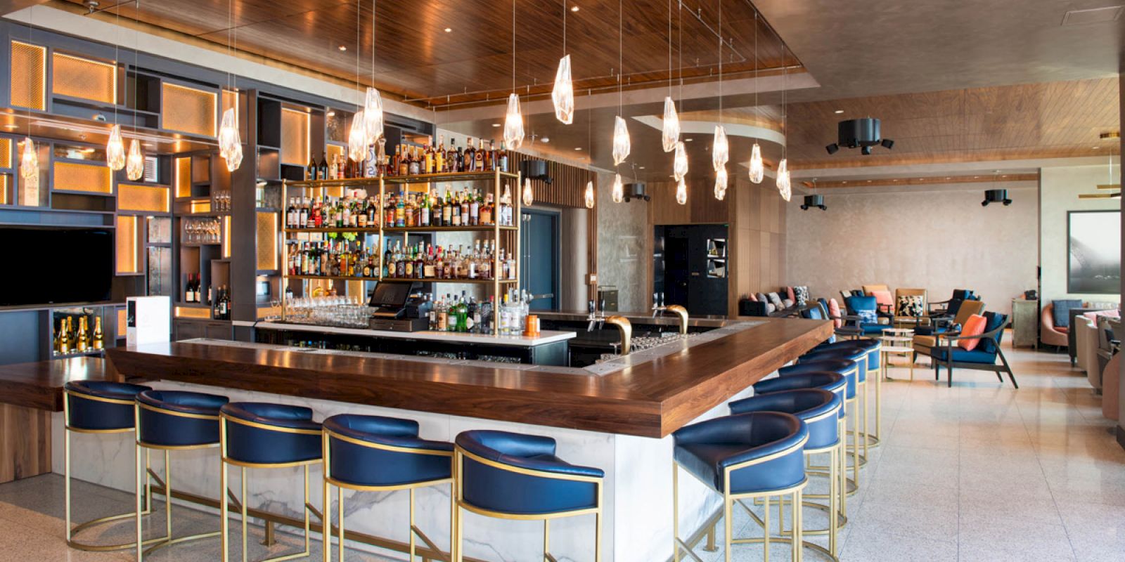 This image shows a modern bar with a central counter, stylish bar stools, hanging lights, and a fully stocked shelf with various bottles of alcohol.