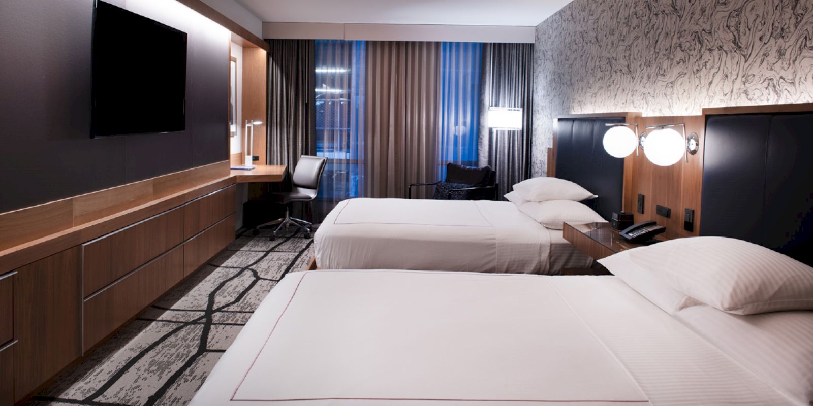 A modern hotel room with two neatly made beds, a wall-mounted TV, a desk, chair, and large windows with curtains.